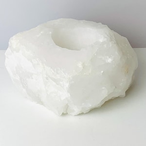 May include: A white, rough-hewn stone candle holder. The natural, irregular shape features a circular indentation to hold a candle. The stone has a crystalline appearance and is set against a white background.
