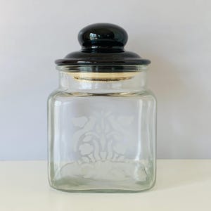 May include: A clear glass jar with a black lid and a floral design etched into the glass. The jar is square shaped and has a wide mouth.