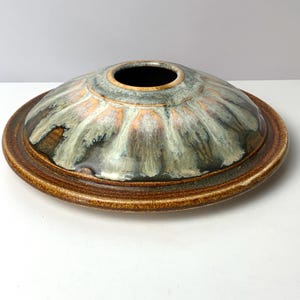 May include: A ceramic bowl with a wide, shallow base and a domed lid. The lid has a central opening and is glazed in shades of green, brown, and gold. The base is a darker brown, creating a contrasting border. The bowl is likely handmade.