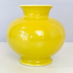 May include: A bright yellow ceramic vase with a rounded, bulbous body, a short neck, and a flared rim. The vase features a smooth, glossy finish that reflects light, and it stands on a circular pedestal base with a clean, solid color.