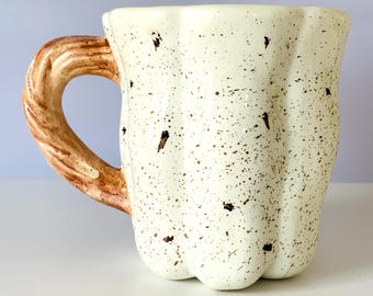 May include: Cream-colored ceramic mug with a speckled design and a brown handle. The mug has a unique, fluted shape and is adorned with dark brown speckles. The handle is a textured, light brown color, adding to the mug's rustic charm.