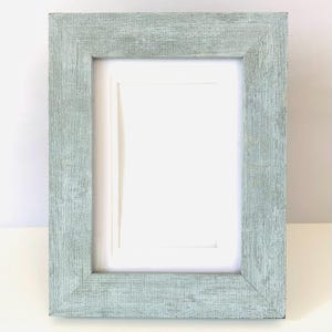 May include: A rectangular picture frame with a light sage green finish and a visible textured wood grain pattern. The frame features a wide border and includes a plain white mat board insert centered within the opening for displaying photos.