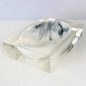 May include: A rectangular clear resin block featuring a recessed oval basin. The interior of the basin displays a marbled pattern with swirling shades of white, gray, and black, creating a high-contrast visual effect within the transparent material.