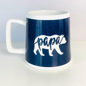 May include: A white ceramic mug with a navy blue exterior and a white interior. The mug features a white bear silhouette with the word "papa" in script. The handle and rim are white.