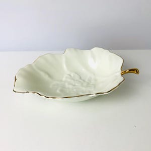 May include: A cream-colored, leaf-shaped ceramic dish with a gold-colored rim and handle. The dish has a textured surface with a grape cluster design in the center. A decorative piece for serving or display.