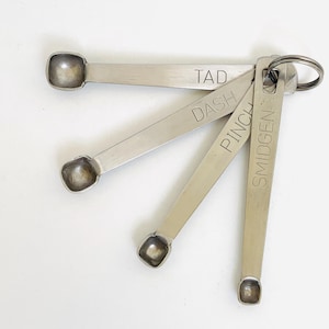 May include: A set of four silver-toned metal measuring spoons held together by a metal ring. Each spoon is engraved with a specific measurement label: Tad, Dash, Pinch, and Smidgen. The spoons feature long, flat handles and small, square-shaped bowls.