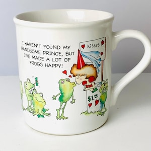May include: White ceramic mug with a whimsical frog and princess design. The mug features the text "I haven't found my handsome prince, but I've made a lot of frogs happy!" and a kissing booth scene with frogs.