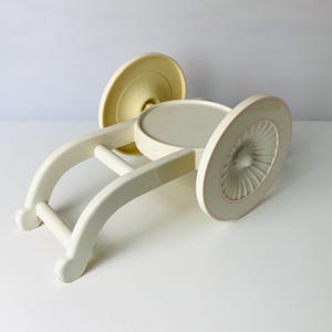 May include: A vintage, off-white decorative stand with a circular top and base, and a curved, arched support structure. The base has a pale yellow color, and the rim of the top has a pink accent. The stand is made of a solid material.