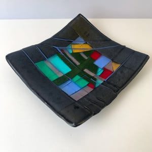 May include: A square, black glass dish with a colorful geometric design. The dish features a dark border and a central mosaic of green, blue, red, yellow, and gray shapes. The dish is a decorative item.