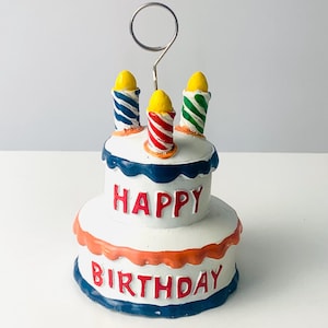 May include: A two-tiered birthday cake-shaped holder with a silver metal clip. The white cake has blue, orange, and red accents, with the words "HAPPY BIRTHDAY" in red. Three colorful candles sit atop the cake.