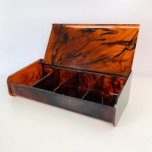 May include: A rectangular storage box with a hinged lid, featuring a glossy tortoiseshell pattern in shades of amber, brown, and black. The interior is divided into three separate compartments by two vertical dividers, providing organized storage space.