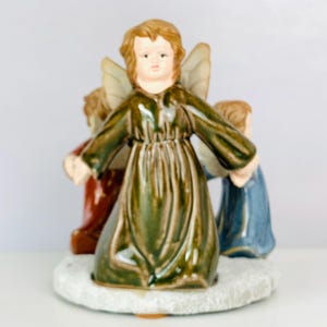 May include: A ceramic figurine of three angels in flowing robes. The central angel wears a green robe, while the others are in red and blue. The angels stand on a white, textured base, with their wings slightly visible. The figurine has a vintage aesthetic.