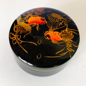 May include: A round, black trinket box with a glossy finish. The lid features a detailed design of orange and gold koi fish and aquatic plants. The box is a decorative item, likely for storing small jewelry or keepsakes.