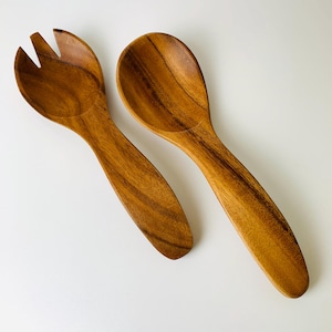 May include: A pair of wooden salad servers. One server is a fork with three tines, and the other is a spoon. Both are made of a light brown wood with visible grain patterns. The servers are designed for serving salads and other dishes.