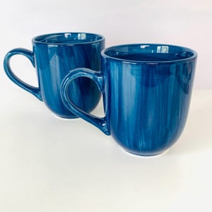 May include: Two ceramic mugs featuring a deep blue glaze with visible vertical brushstroke textures. Each mug has a rounded body, a curved handle on the side, and a smooth, glossy finish. The mugs are positioned side by side against a plain background.