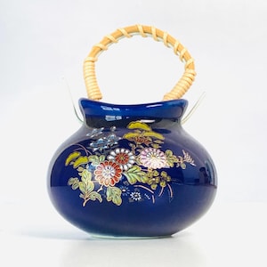 May include: A cobalt blue ceramic vase with a woven handle. The vase features a floral design in red, white, pink, and green. The handle is made of woven light brown material.