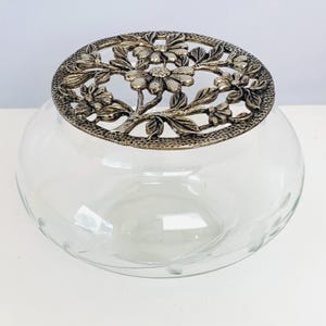 May include: A clear glass bowl with a silver-colored floral design lid. The bowl has a round shape and etched floral details. The lid features an intricate pattern of flowers and leaves, adding a decorative touch to the glass bowl.