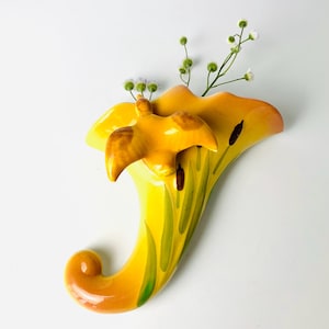 May include: A yellow ceramic wall vase with a bird accent. The vase has a curved shape with green and brown painted details. Small white flowers and green stems are placed inside the vase.