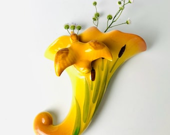 May include: A yellow ceramic wall vase with a bird accent. The vase has a curved shape with green and brown painted details. Small white flowers and green stems are placed inside the vase.