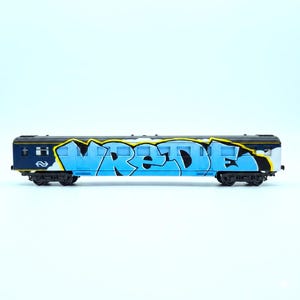 Pode incluir: Um vagão de trem modelo azul e preto com arte de grafite. A palavra "VREDE" está escrita em azul claro e preto com contornos amarelos na lateral do vagão.