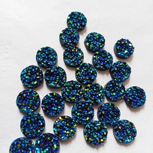 以下が含まれることがあります： 虹色の青と緑のグリッターが付いた丸いカボションが20個セットです。カボションは直径約2.5cmで、裏側は平らです。