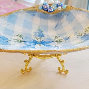May include: A decorative seashell-shaped dish with a blue and white gingham pattern and floral accents. The dish is supported by a gold-colored metal stand with ornate legs. The rim is trimmed with gold and adorned with beads.