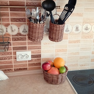 May include: Two brown wicker baskets hanging on a tiled wall, each holding a variety of kitchen utensils. The baskets are hung from a metal rod. A bowl of fruit sits on a black countertop below the baskets.