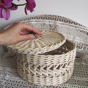 May include: A white wicker basket with a lid. The basket is round and has a woven pattern. The lid is slightly ajar and the basket is sitting on a white lace tablecloth.