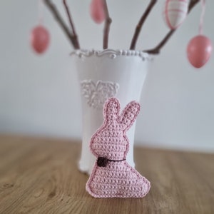 May include: A pink crocheted bunny rabbit with a brown bow. The bunny is sitting on a wooden surface in front of a white vase with a floral design.
