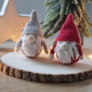 Könnte beinhalten: Zwei gehäkelte Gnome, einer grau und einer rot, mit weißen Bärten, die auf einer Holzscheibe sitzen.