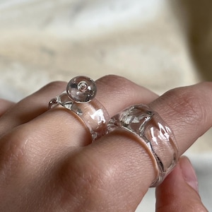 Puede incluir: Dos anillos de vidrio transparente. Un anillo presenta una pequeña cuenta redonda de vidrio transparente en la parte superior. El otro anillo es una banda gruesa de vidrio transparente. Ambos anillos están en los dedos de una persona.