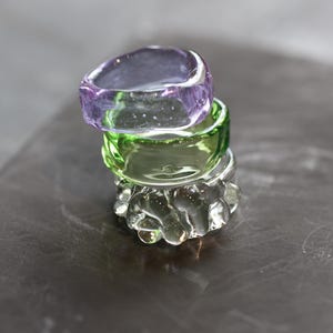 Può includere: Una pila di tre anelli di vetro nei toni del viola, verde e trasparente. L'anello superiore è viola traslucido, l'anello centrale è verde brillante e l'anello inferiore è trasparente con un design strutturato e a bolle. Gli anelli sono su una superficie scura.