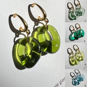 Könnte beinhalten: Goldene Creolen-Ohrringe mit olivgrünen, ovalen Glasanhängern. Das Bild zeigt auch vier weitere Ohrringpaare in Grün- und Blautönen. Die Ohrringe haben ein modernes Design.