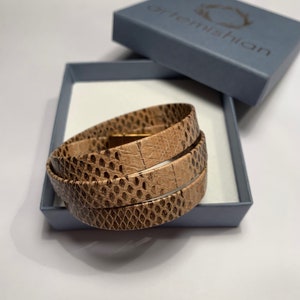 Könnte beinhalten: Ein mehrfach gewickeltes Armband mit einem beigen und braunen Schlangenledermuster und einem metallisch goldfarbenen Verschluss. Das Armband liegt in einer hellblauen Schachtel mit dem Schriftzug artemishian auf dem Deckel.