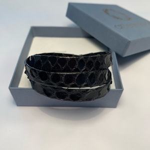 Op de afbeelding: Twee zwarte leren armbanden met een reptielschubbenpatroon worden gepresenteerd in een lichtblauwe doos. Het deksel van de doos is open en toont de armbanden. De armbanden zijn opgerold. De doos staat op een wit oppervlak.
