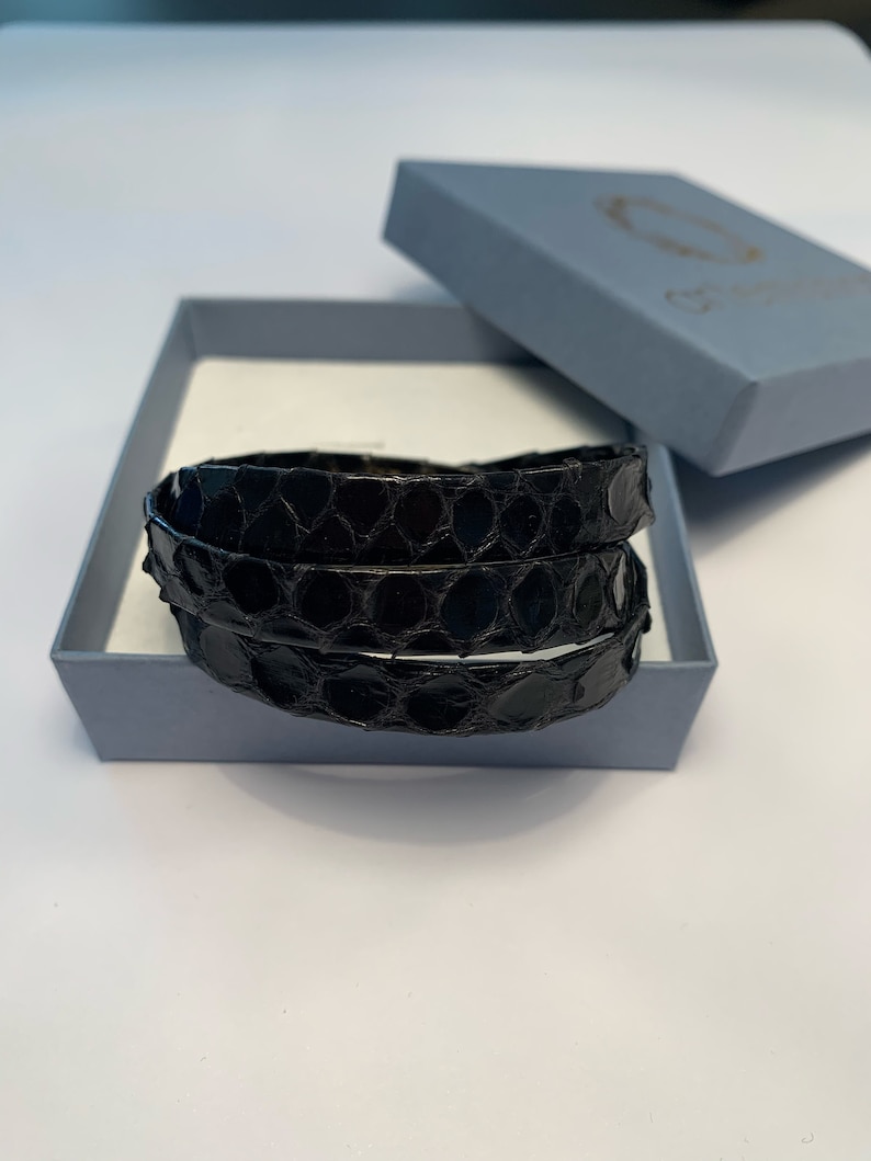 Op de afbeelding: Twee zwarte leren armbanden met een reptielschubbenpatroon worden gepresenteerd in een lichtblauwe doos. Het deksel van de doos is open en toont de armbanden. De armbanden zijn opgerold. De doos staat op een wit oppervlak.