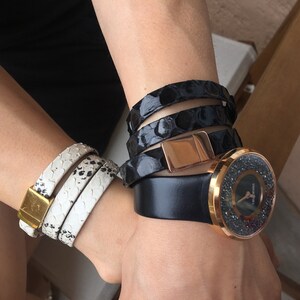 Op de afbeelding: Een pols versierd met een verzameling accessoires. Een wit en zwart slangenprint wikkelarmband met een gouden sluiting, een zwarte wikkelarmband met een ros&eacute;gouden sluiting en een zwart horloge met een met kristallen bezette lunette.