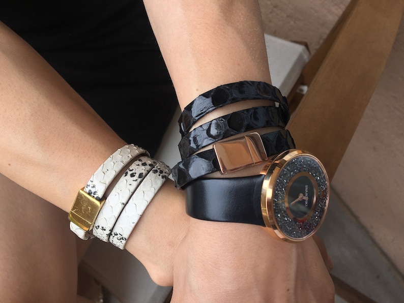 Op de afbeelding: Een pols versierd met een verzameling accessoires. Een wit en zwart slangenprint wikkelarmband met een gouden sluiting, een zwarte wikkelarmband met een ros&eacute;gouden sluiting en een zwart horloge met een met kristallen bezette lunette.