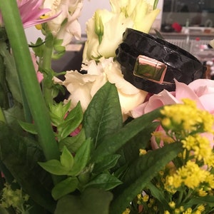 Op de afbeelding: Zwarte leren armband met een goudkleurige sluiting, rustend tussen een boeket bloemen. De armband heeft een textuur, slangenleerachtig patroon. De bloemen omvatten witte, roze en gele bloemen met groene bladeren.