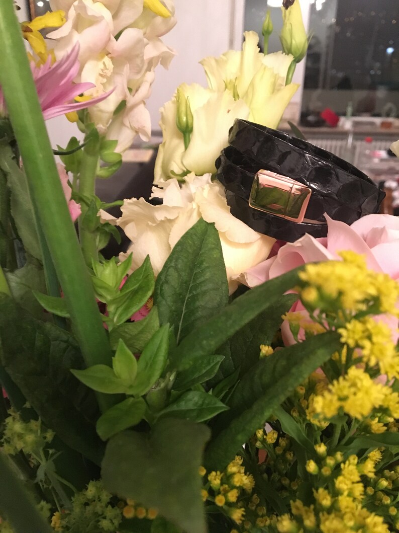 Op de afbeelding: Zwarte leren armband met een goudkleurige sluiting, rustend tussen een boeket bloemen. De armband heeft een textuur, slangenleerachtig patroon. De bloemen omvatten witte, roze en gele bloemen met groene bladeren.