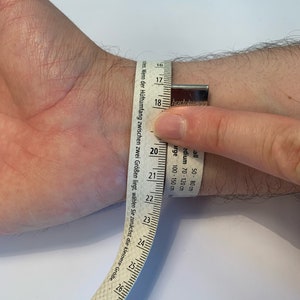 Op de afbeelding: Een pols wordt gemeten met een witte meetlint. De tape toont metingen in centimeters, waarbij de pols ongeveer 18 cm meet. De tape heeft ook tekst in het Duits.