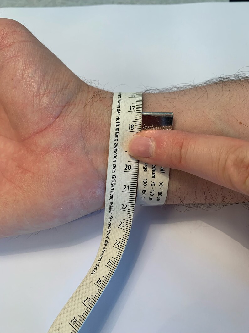 Op de afbeelding: Een pols wordt gemeten met een witte meetlint. De tape toont metingen in centimeters, waarbij de pols ongeveer 18 cm meet. De tape heeft ook tekst in het Duits.