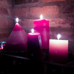 May include: Four pink candles, two are pyramid shaped, one is square, and one is rectangular, all lit and burning.