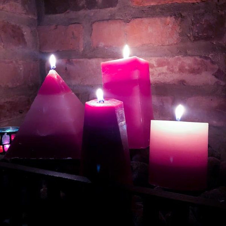 May include: Four pink candles, two are pyramid shaped, one is square, and one is rectangular, all lit and burning.