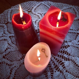 May include: Three red candles with white accents are lit and burning on a white crocheted doily. The candles are in different shapes and sizes. One candle has a small key charm on top.