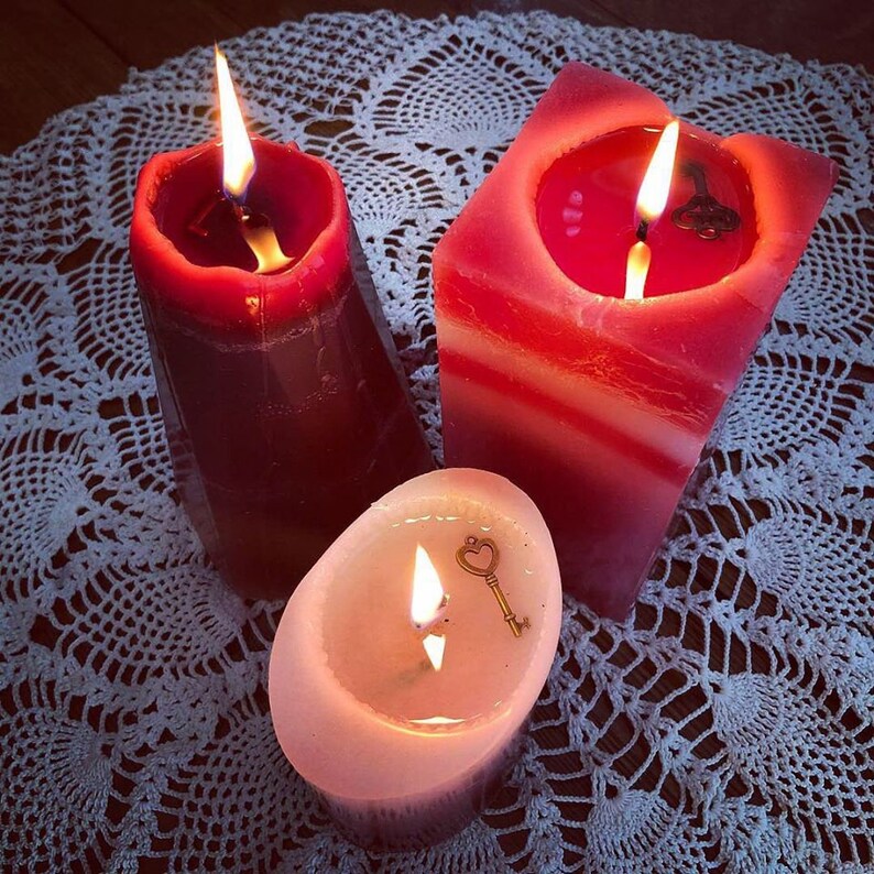 May include: Three red candles with white accents are lit and burning on a white crocheted doily. The candles are in different shapes and sizes. One candle has a small key charm on top.