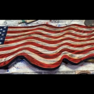 May include: A wooden American flag with a distressed finish. The flag features the traditional red and white stripes and a blue field with white stars. The edges of the flag have a wavy design.