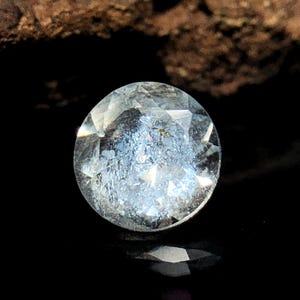 May include: A clear, faceted gemstone with a light blue hue. The gemstone is round and has a reflective surface.