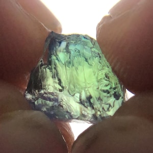 May include: A green and blue gemstone with a faceted cut. The gemstone is held between two fingers.