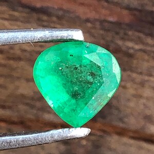May include: A pear-shaped emerald gemstone, held by tweezers, showcasing a deep green hue. The gem's facets are visible, reflecting light and revealing internal inclusions. The close-up view emphasizes the emerald's natural characteristics and the quality of the cut.